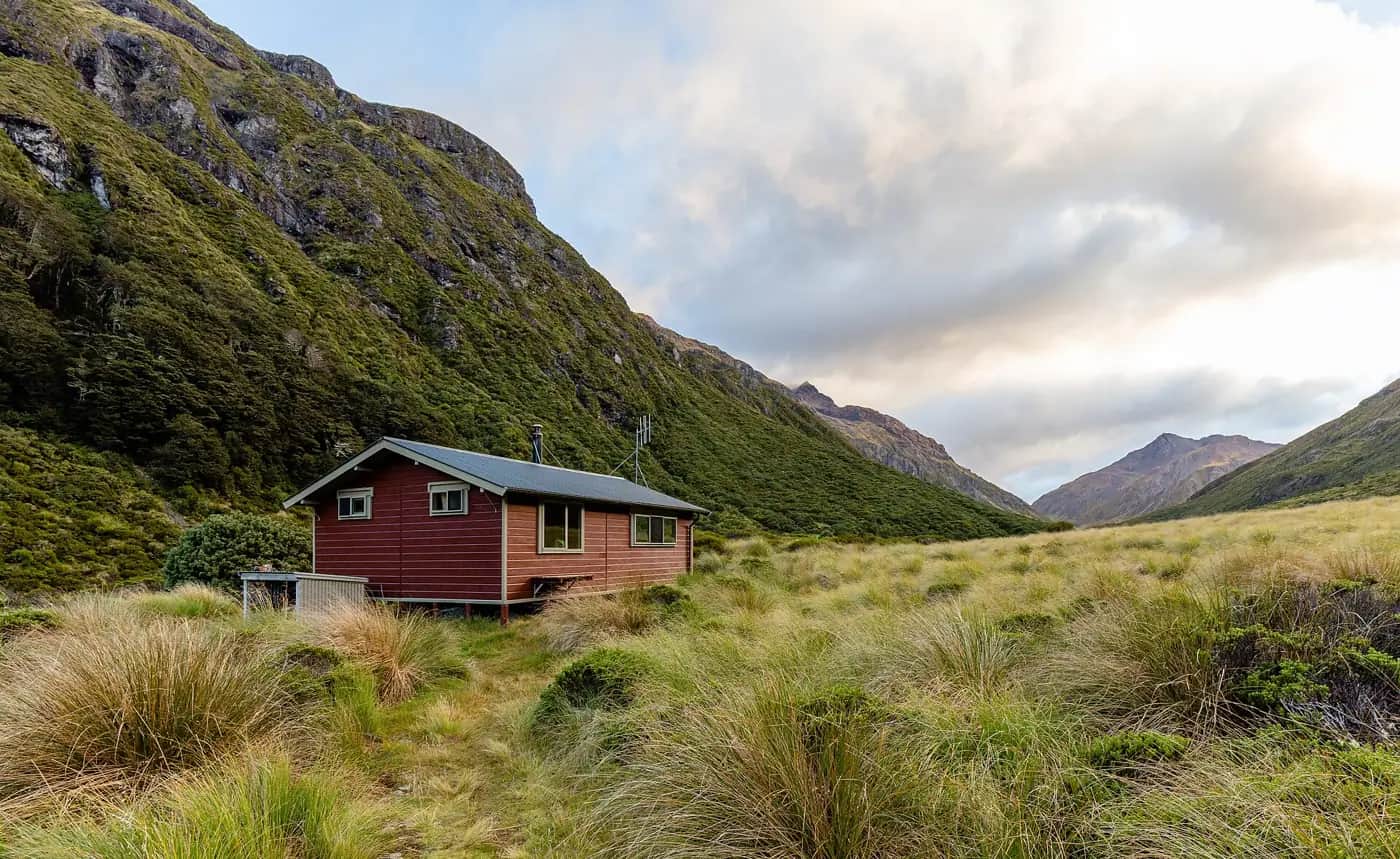 A small red wooden tramping hut in a tussock valley below steep green mountainsides, with cloud building over the peaks
