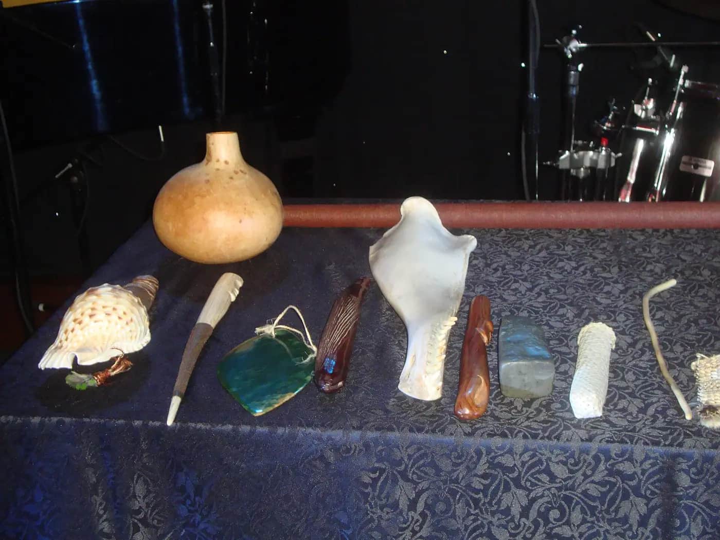 A table covered with a dark patterned cloth, laid out with taonga pūoro: a large gourd, a spotted conch shell, a carved flute, a green stone piece on a cord, a bone and several carved wooden pieces, with a drum kit on the stage behind