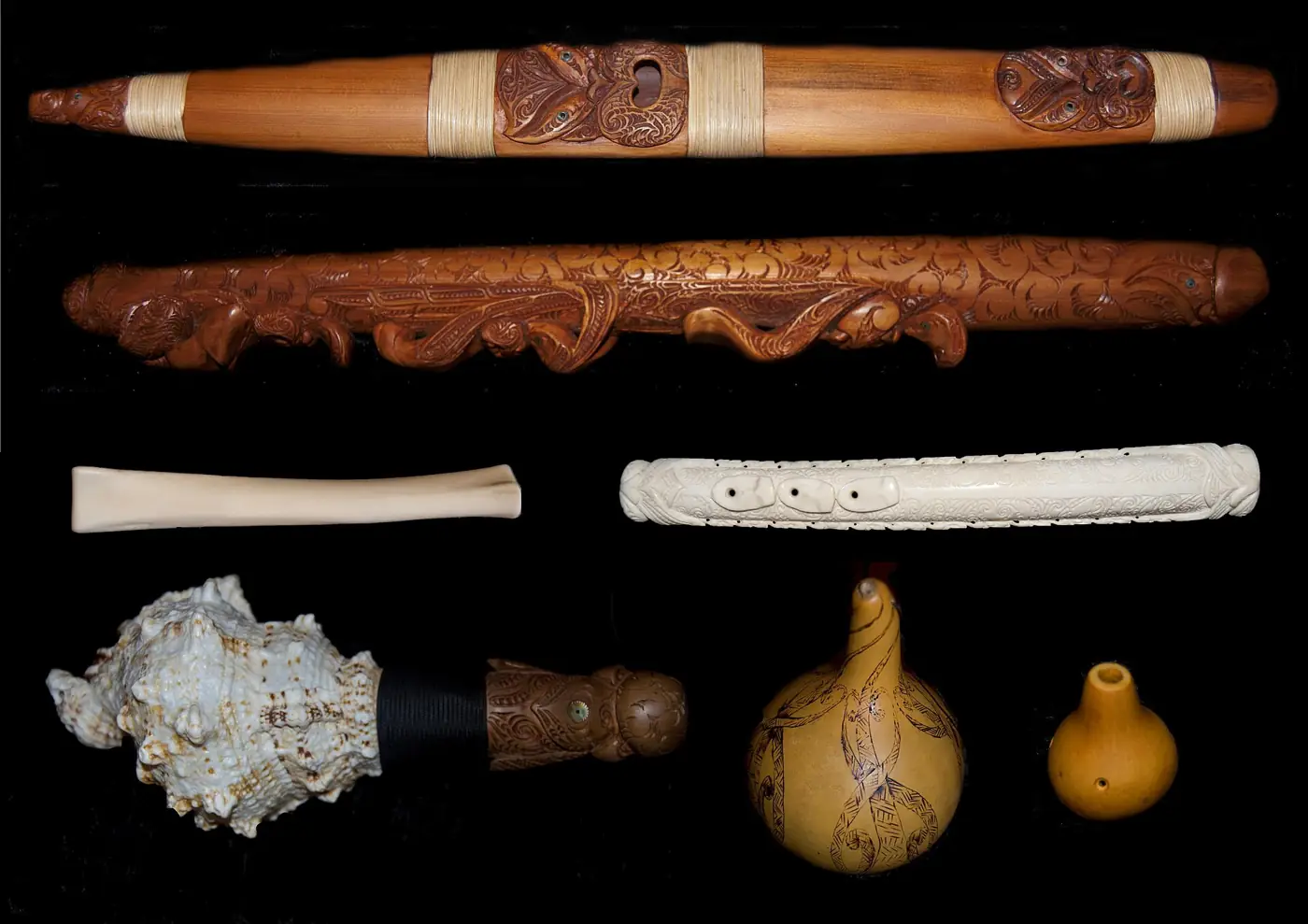 Taonga pūoro laid out on a black background: two long carved wooden flutes bound with fibre, a plain bone flute, a carved white flute, a spiky conch shell trumpet with a carved wooden mouthpiece, and two gourd instruments
