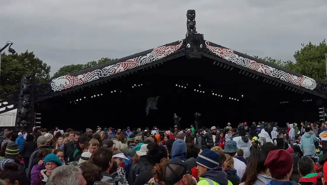A huge crowd gathered in front of a large black stage shaped like a wharenui, with red and white painted patterns along its roof and a carved figure on top