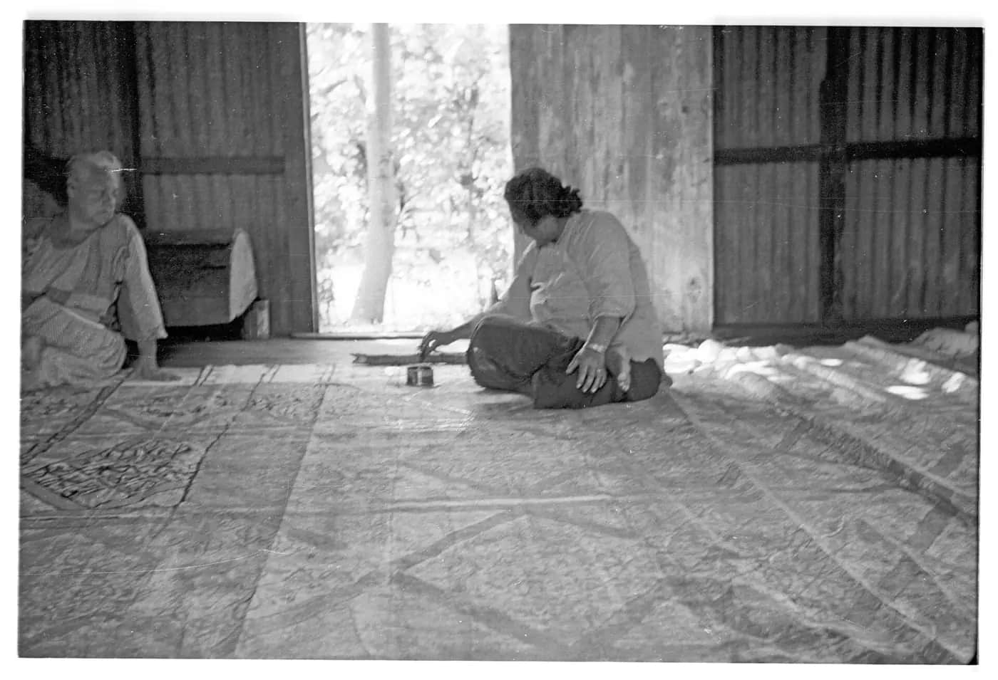 Black-and-white photo of two women seated on the floor of a wooden house beside a very large sheet of patterned barkcloth