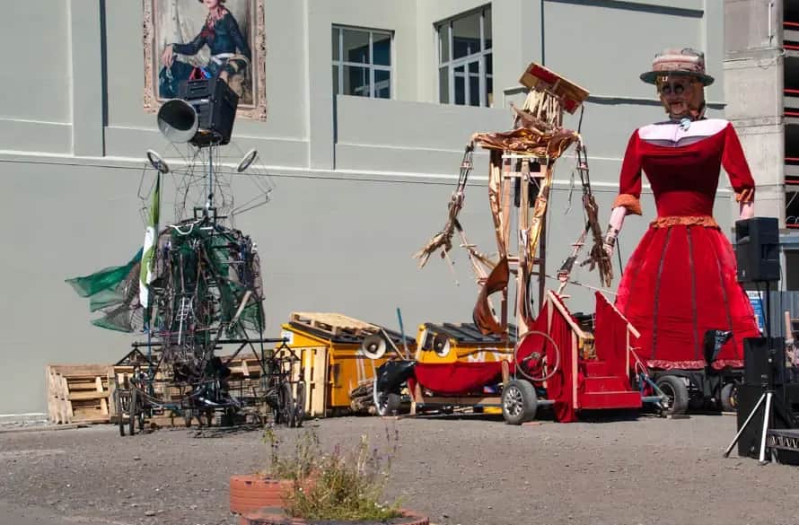 Giant street-theatre puppets outside a building: a tall woman in a red dress and hat, a wooden skeleton-like figure and a wire creature on wheels, with speakers beside them