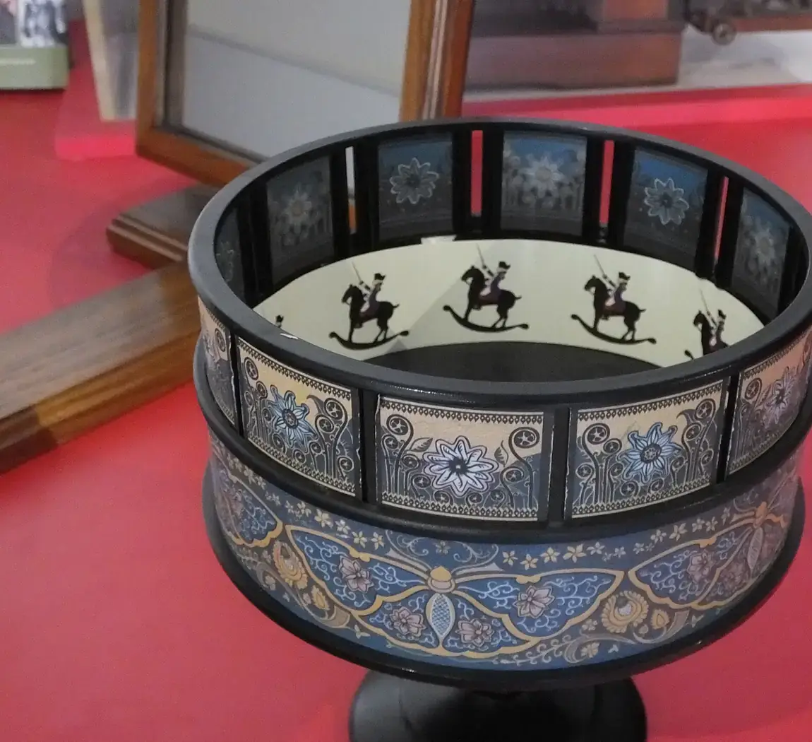 A round drum-shaped zoetrope with slits around the top and a strip of pictures of a figure on a rocking horse inside, on a red museum table