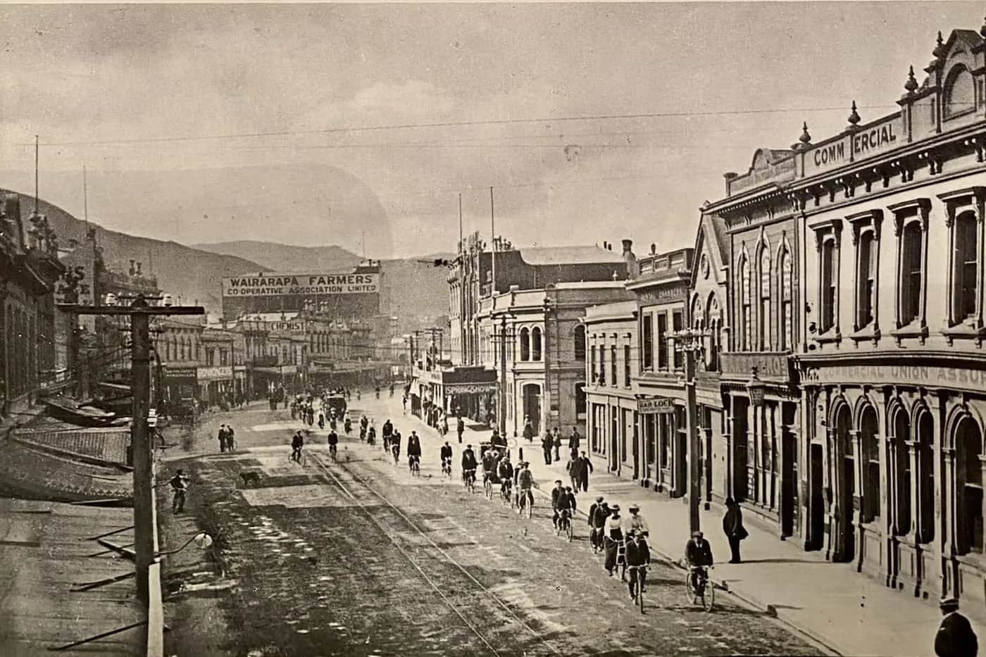 Old black and white photo of a busy Wellington street around 1902, full of cyclists, with Victorian shopfronts and hills behind