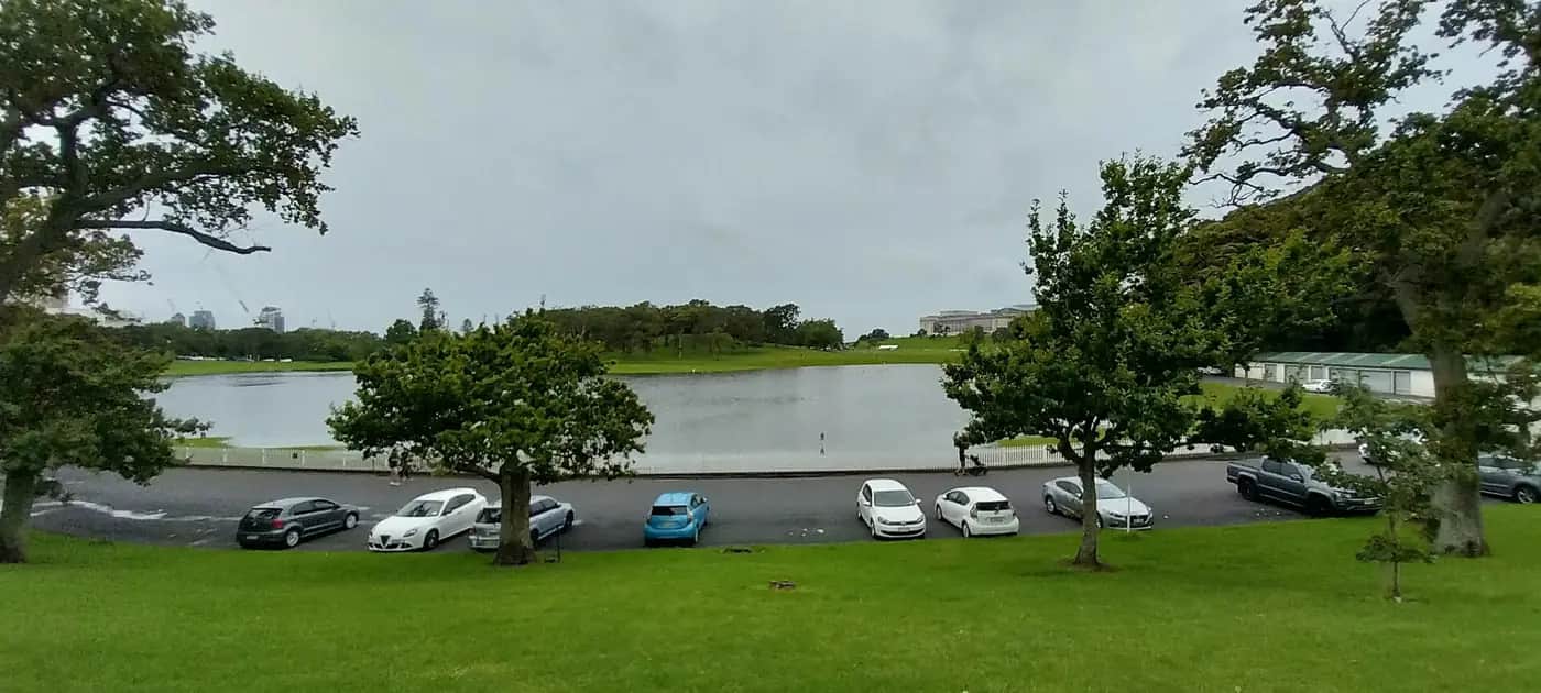 Floodwater forming a lake over the cricket fields in the Auckland Domain, with parked cars and trees in the foreground