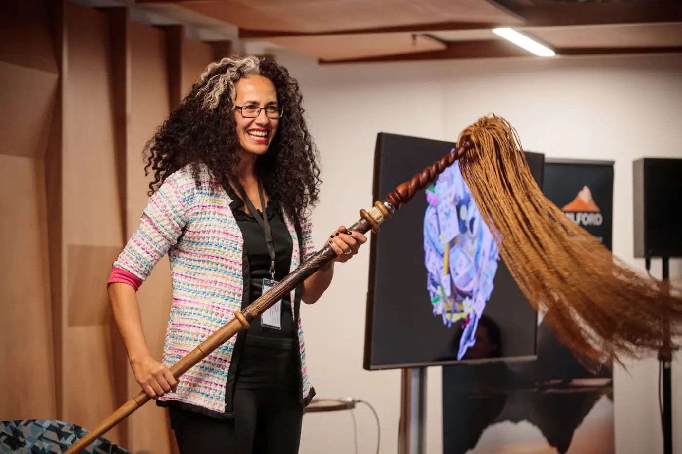 Poet Selina Tusitala Marsh smiling on stage, holding a long carved wooden staff topped with a flowing fibre tassel