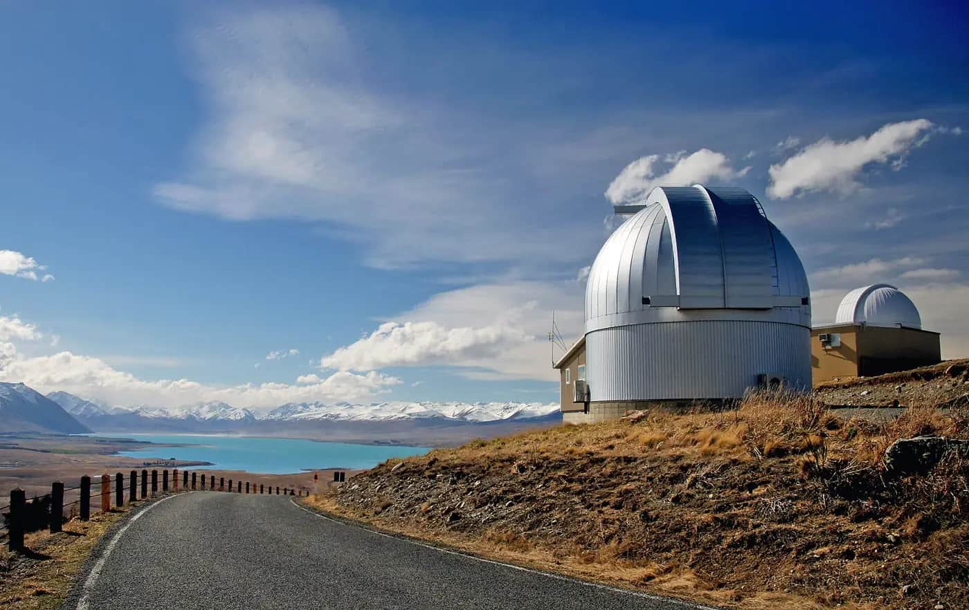 A silver observatory dome on a dry hilltop above a turquoise lake and snowy mountains under a blue sky