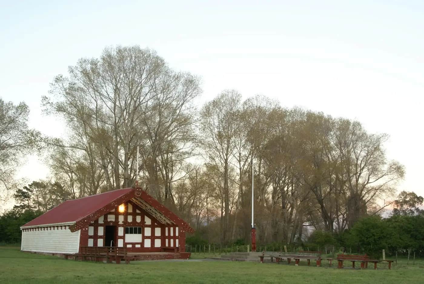 The carved red and white wharenui Hinetapora at Mangahanea Marae at dusk, with benches facing the open marae ātea
