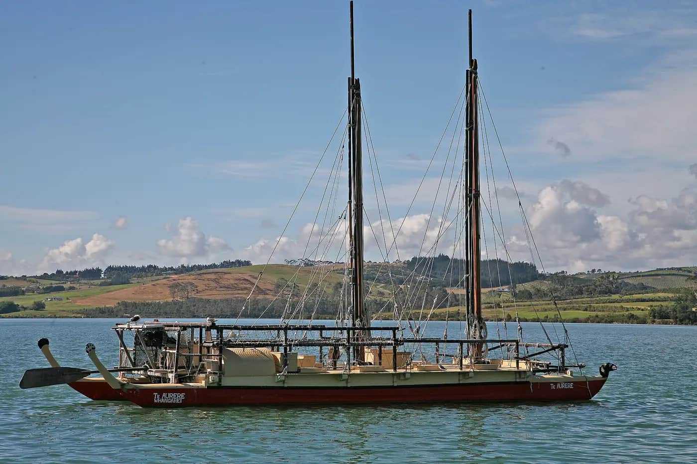 Te Aurere, a red-hulled double-hulled voyaging waka with two tall masts, anchored in a calm harbour with green farmland behind