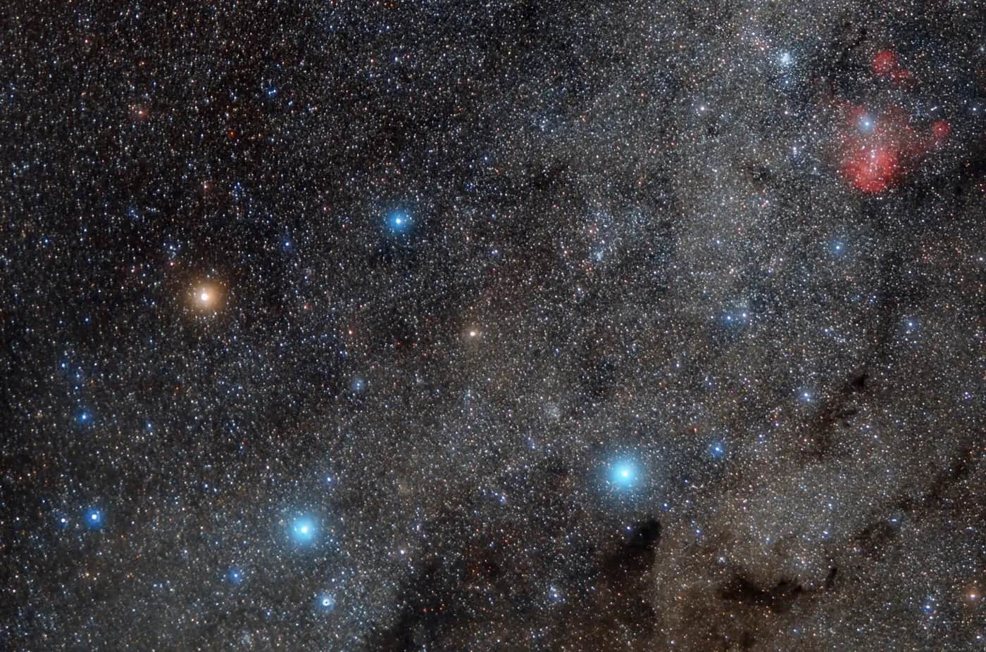 A dense star field with the four bright blue-white stars of the Southern Cross, one orange star, and a red glowing nebula
