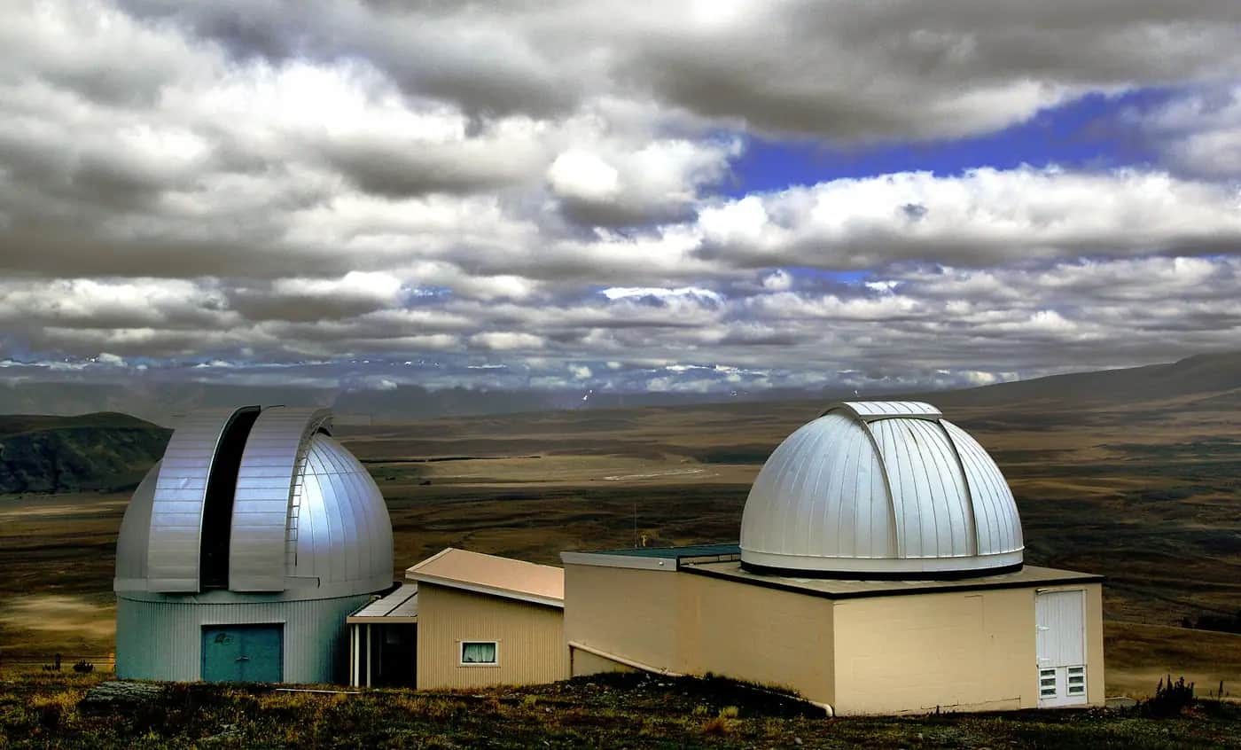 Two silver telescope domes on a brown hilltop at Mount John University Observatory under a cloudy sky
