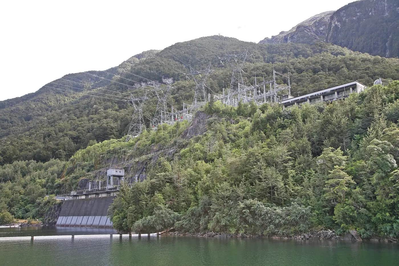 Manapōuri power station switchyard with pylons and power lines climbing a bush-covered hillside above Lake Manapōuri