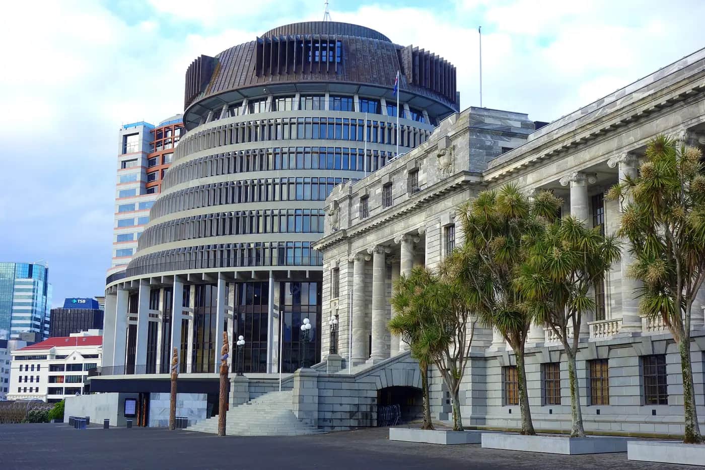 The round Beehive building and the grey stone Parliament House in Wellington, with cabbage trees and carved pou in front