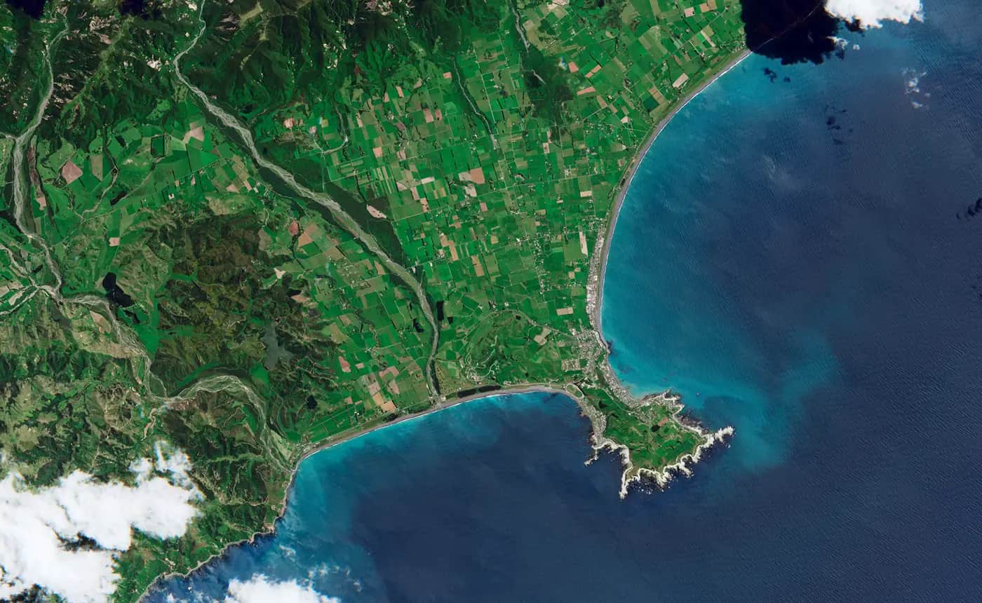 Satellite view of the Kaikōura Peninsula and farmland beside a blue sea, with pale new rocky shoreline around the coast