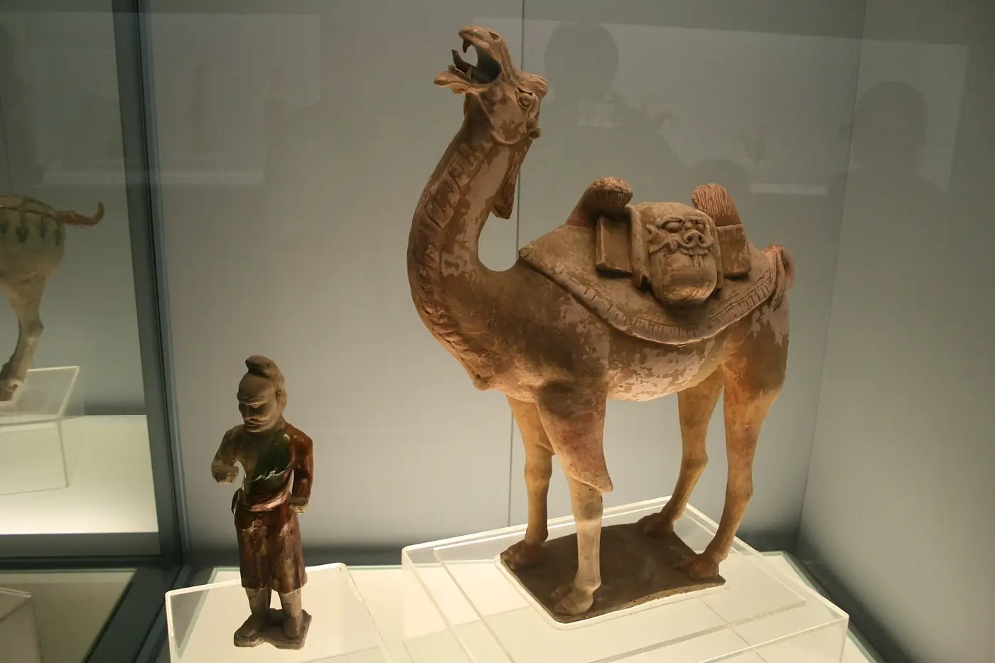A pottery figure of a two-humped camel with a saddle and packs, next to a small figure of a bearded camel driver in a museum case