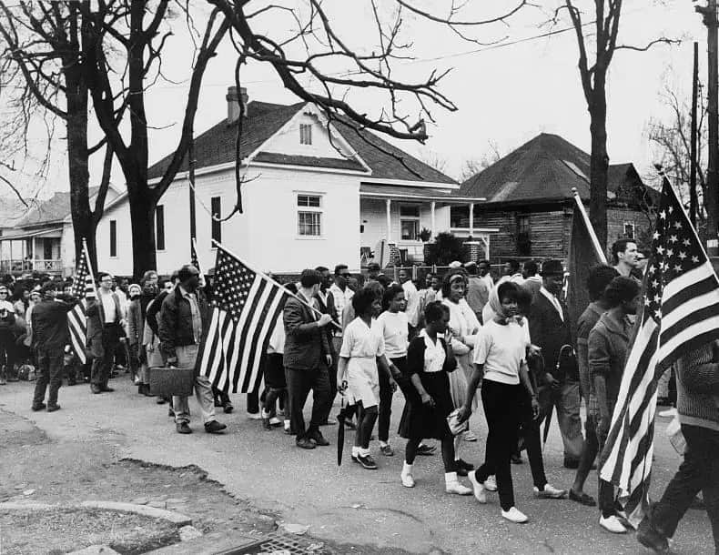 Black and white photograph of marchers, many of them young, walking along a street carrying American flags past wooden houses