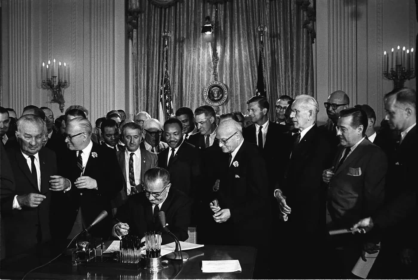 Black and white photograph of President Lyndon Johnson signing a law at a desk surrounded by a crowd of men in suits, including Martin Luther King Jr