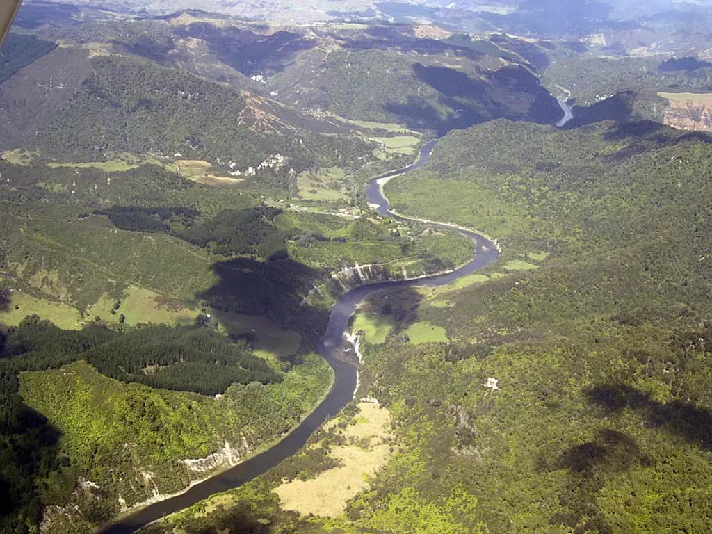 Aerial view of the Whanganui River curving through steep bush-covered hills and pale cliffs, with small farms on the flat river terraces