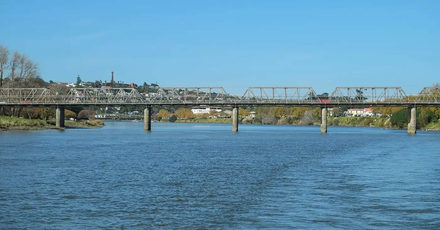 A wide blue river in a town, crossed by a long steel truss bridge, with houses and a tower on a hill behind