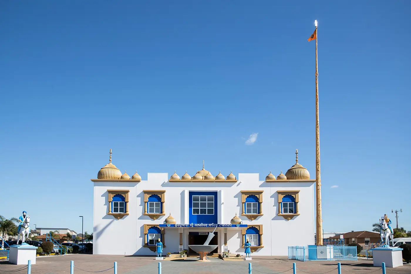 A large white two-storey gurdwara with golden domes and blue windows under a clear sky, with a tall flagpole and statues of riders on horseback at each side