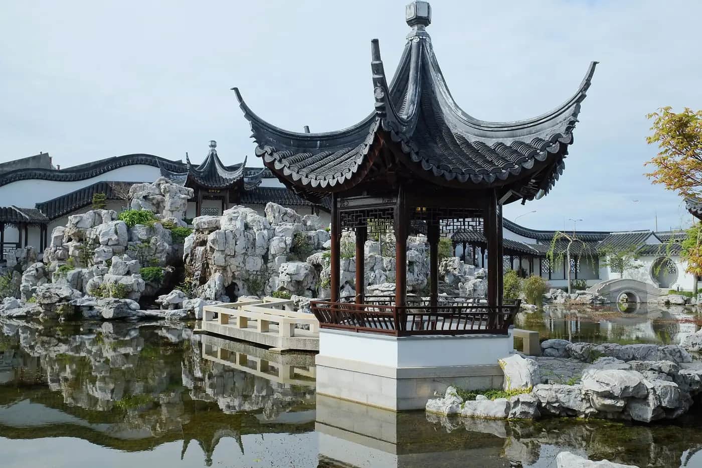 A Chinese pavilion with curved tiled roofs standing in a still pond, surrounded by grey limestone rocks and white walls