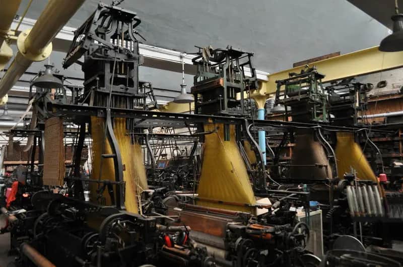 Rows of old Jacquard looms in a textile mill, with punched-card chains hanging above the golden threads.