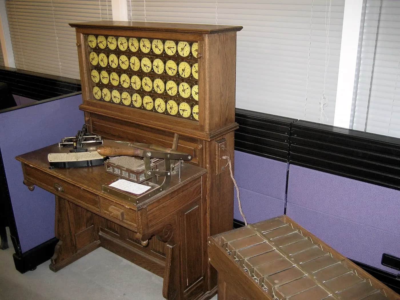 A wooden Hollerith tabulating machine from the 1890s, with rows of round counting dials above a desk holding a punched-card press.