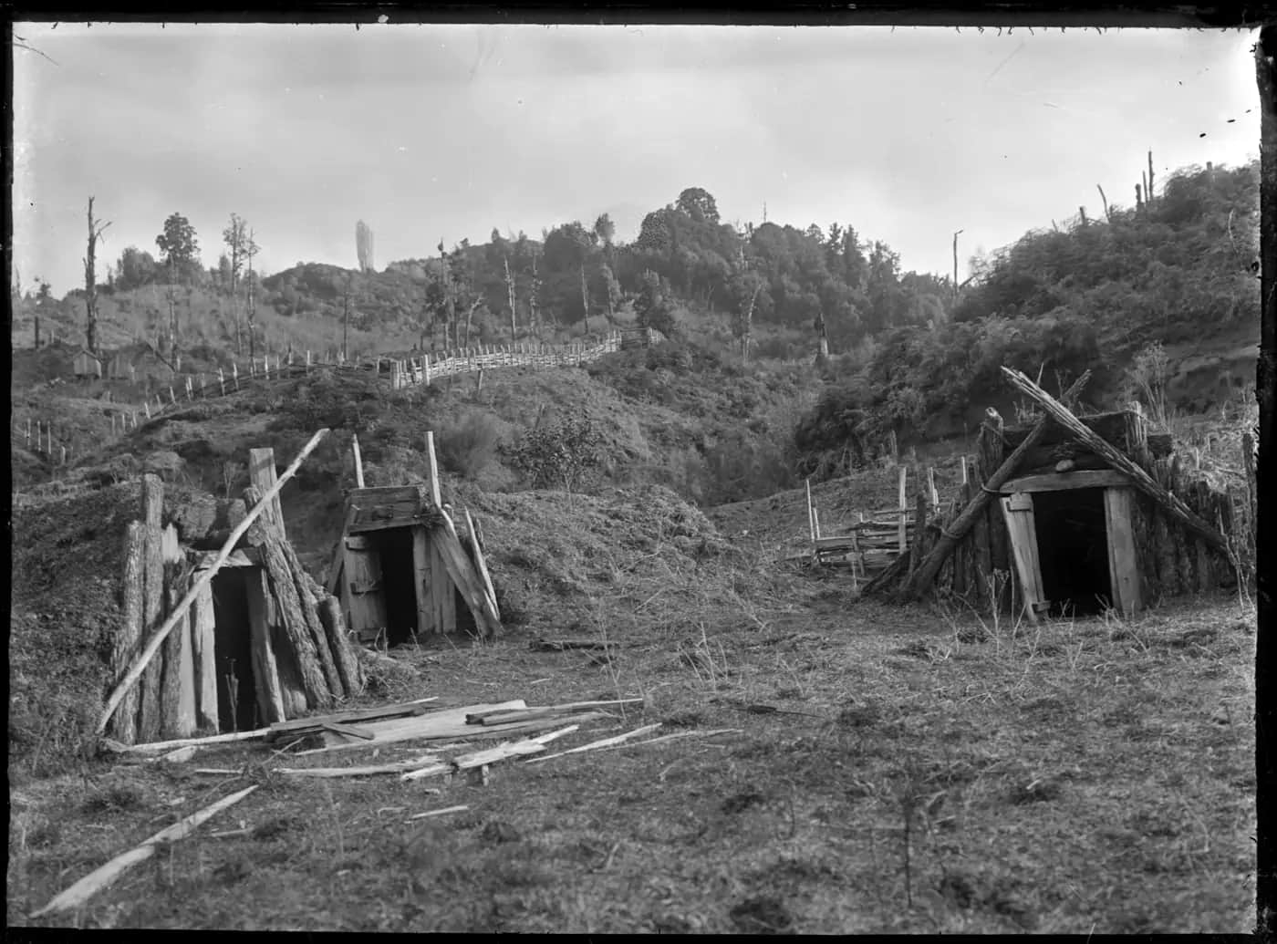 Black and white photo of three timber-framed kūmara storage pits dug into a hillside