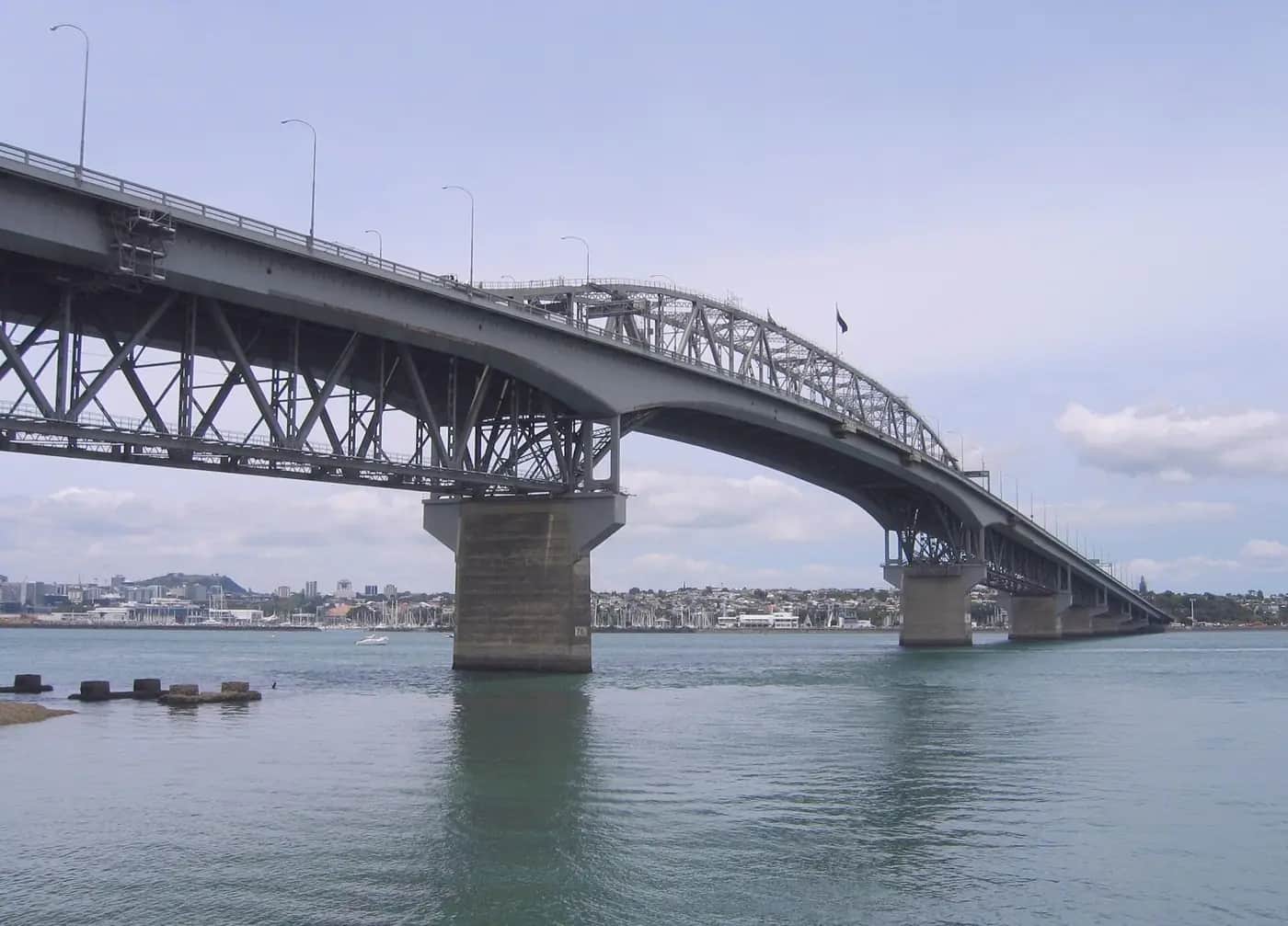The Auckland Harbour Bridge seen from below, showing its steel truss frame made of triangles resting on concrete piers.
